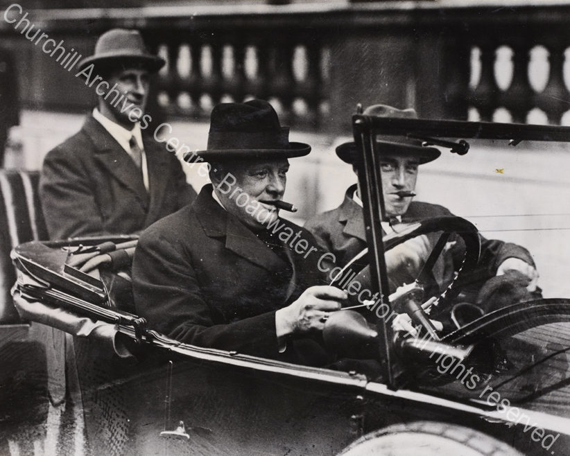 Photo shows WSC wearing a hat at the wheel of an open topped car with Cunliffe-Lister in the passenger seat. Both have cigars in their mouths.