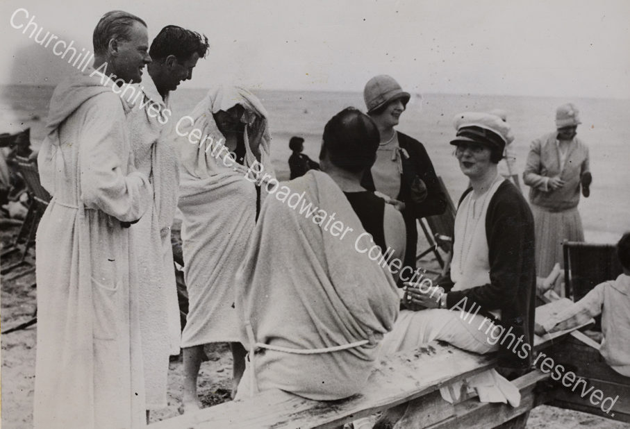 Beach scene with a group of people in bathing robes. One (?WSC) has his head concealed with a robe.