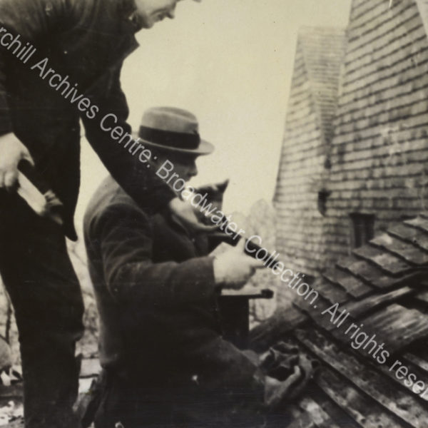 Photo shows WSC kneeling on the roof [at Chartwell] being handed a roof tile by a workman. WSC lifts a cigar to his mouth.