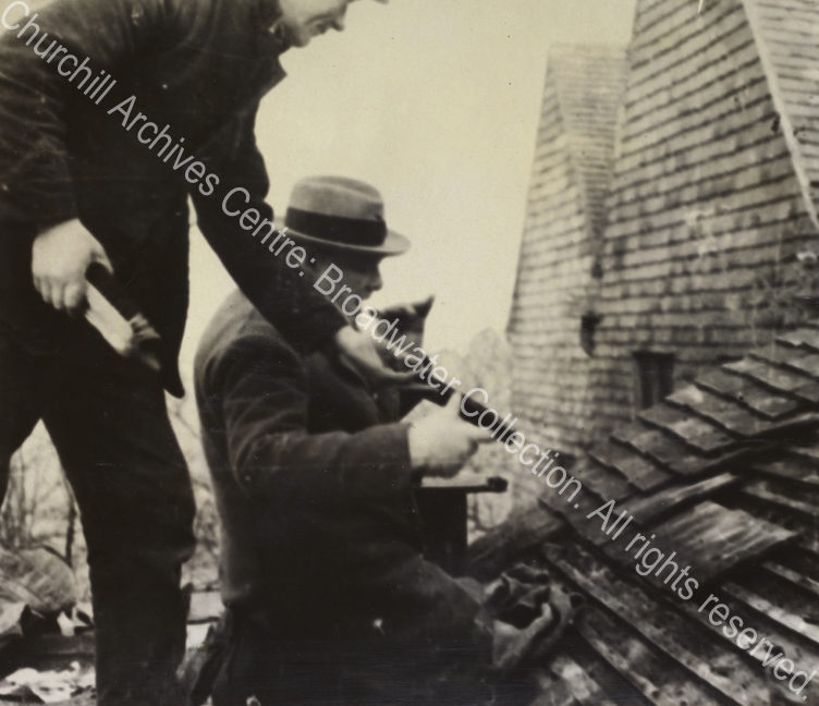 Photo shows WSC kneeling on the roof [at Chartwell] being handed a roof tile by a workman. WSC lifts a cigar to his mouth.