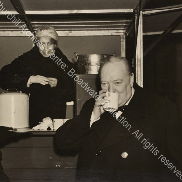 Photo shows WSC drinking from a mug. A female ?canteen worker stands behind him.