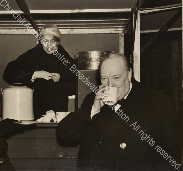 Photo shows WSC drinking from a mug. A female ?canteen worker stands behind him.
