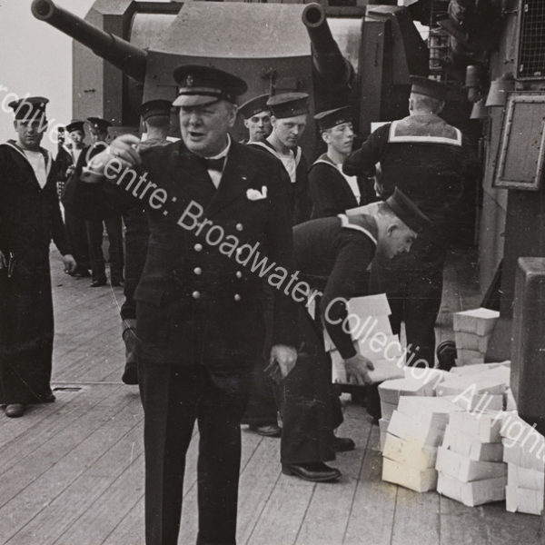 Photo shows WSC on the deck of HMS Prince of Wales. He is wearing a double-breasted jacket and a peaked cap and gesticulating. Behind him are Royal Navy sailors in uniform working to carry piles of boxes [food parcels given to each man as a gift from President Roosevelt].