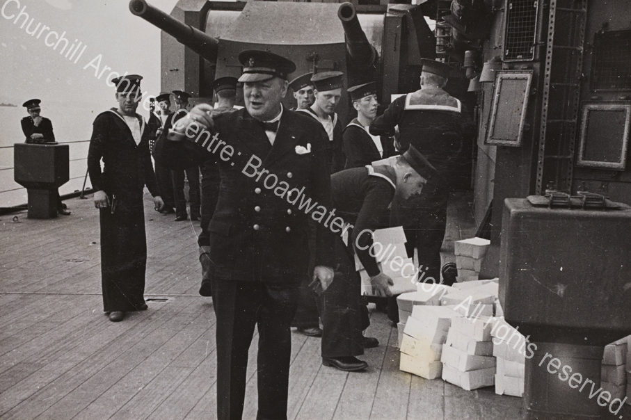 Photo shows WSC on the deck of HMS Prince of Wales. He is wearing a double-breasted jacket and a peaked cap and gesticulating. Behind him are Royal Navy sailors in uniform working to carry piles of boxes [food parcels given to each man as a gift from President Roosevelt].
