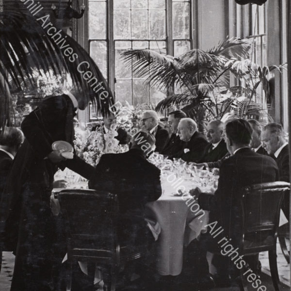 Photo shows a formal gathering [Allied ambassadors’ lunch at the Soviet embassy] seated at table amid large potted ferns. WSC is in the centre listening to talk at the table. Seated on his left is Ivan Maisky