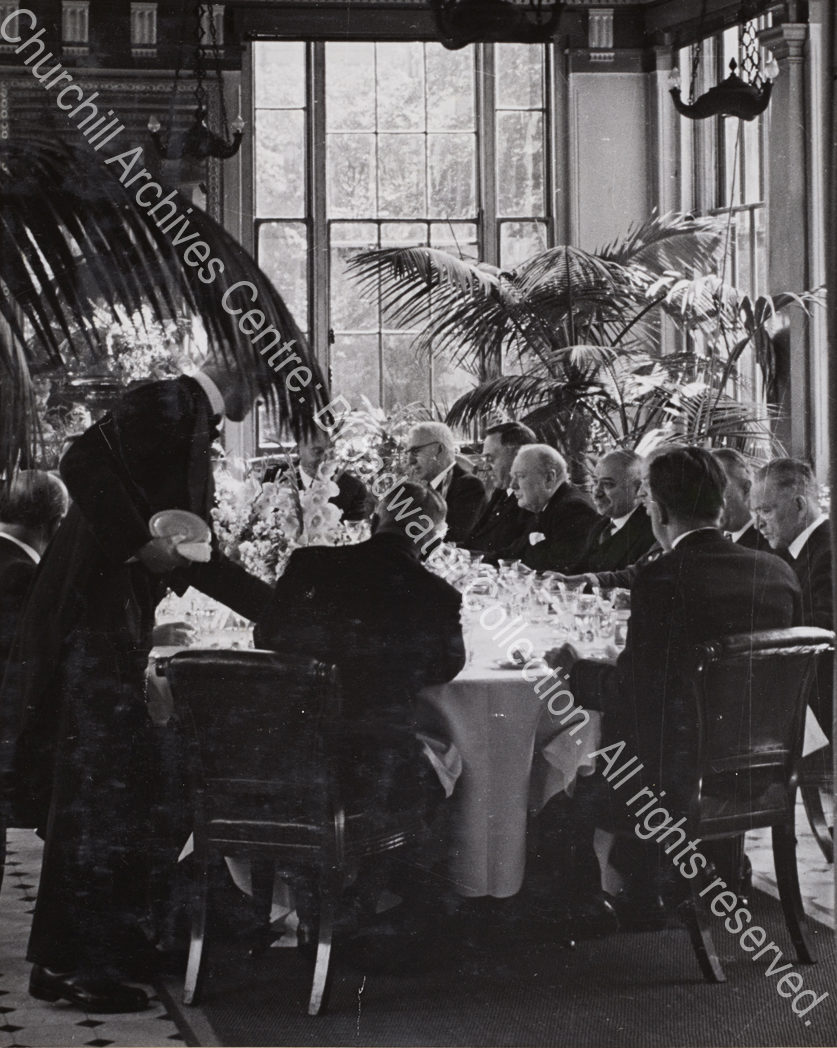 Photo shows a formal gathering [Allied ambassadors’ lunch at the Soviet embassy] seated at table amid large potted ferns. WSC is in the centre listening to talk at the table. Seated on his left is Ivan Maisky
