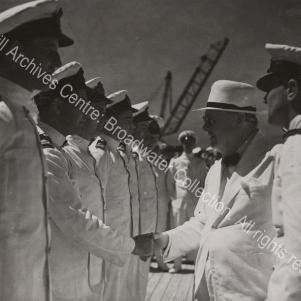 Photo shows WSC smiling as he shakes hands with a man wearing a white naval uniform. A line of men in white naval uniforms and caps stand in front of WSC.