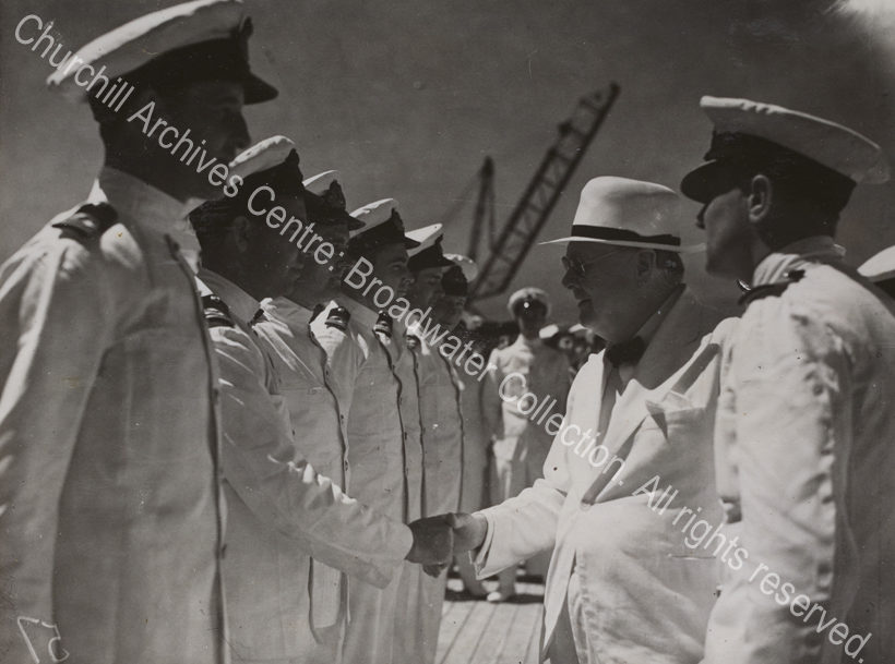 Photo shows WSC smiling as he shakes hands with a man wearing a white naval uniform. A line of men in white naval uniforms and caps stand in front of WSC.