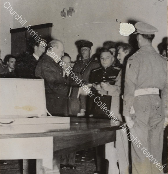 Photo shows WSC handing the sword to Stalin as a tribute at the Soviet Legation in Tehran [Iran]. Anthony Eden [later 1st Lord Avon] is just seen behind WSC and they are watched by a group of people.