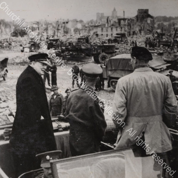 Photo shows WSC standing in the back of an army jeep (wearing an overcoat and peaked cap with a cigar in his mouth) in conversation with General Bernard Montgomery (wearing a beret and facing away from the camera). In the background are the ruins of Caen plus many army vehicles and military personnel.