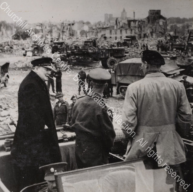 Photo shows WSC standing in the back of an army jeep (wearing an overcoat and peaked cap with a cigar in his mouth) in conversation with General Bernard Montgomery (wearing a beret and facing away from the camera). In the background are the ruins of Caen plus many army vehicles and military personnel.