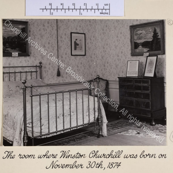 Photo shows interior view of bedroom with bed and chest of drawers. Items relating to WSC including framed facsimiles of documents and paintings by WSC are on display.