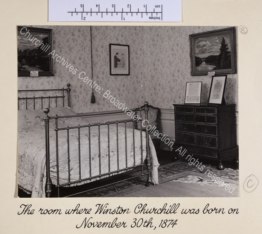 Photo shows interior view of bedroom with bed and chest of drawers. Items relating to WSC including framed facsimiles of documents and paintings by WSC are on display.