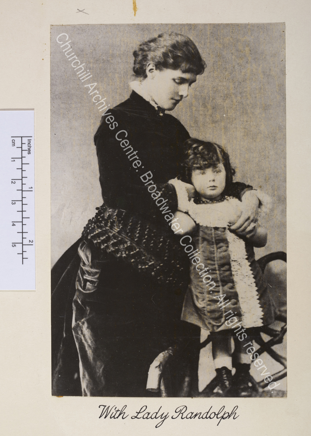 Photograph showing WSC as a toddler standing on a chair leaning against his mother. WSC has long curly hair and is wearing a velvet and lace dress