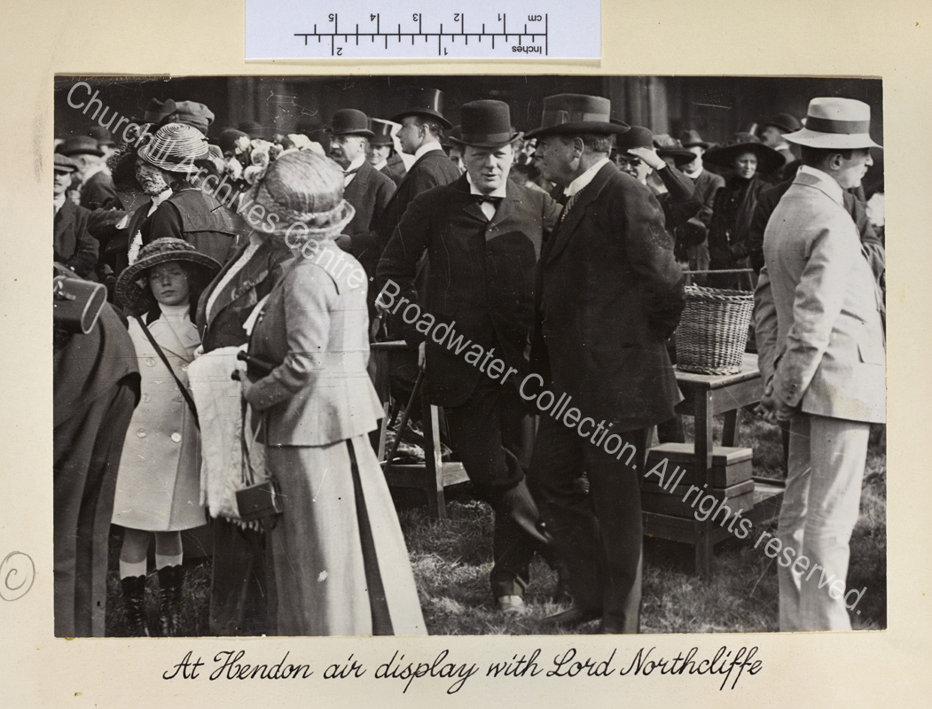 WSC leaning on a stick talking to Lord Northcliffe [formerly Alfred Harmsworth] in a crowd of people.
