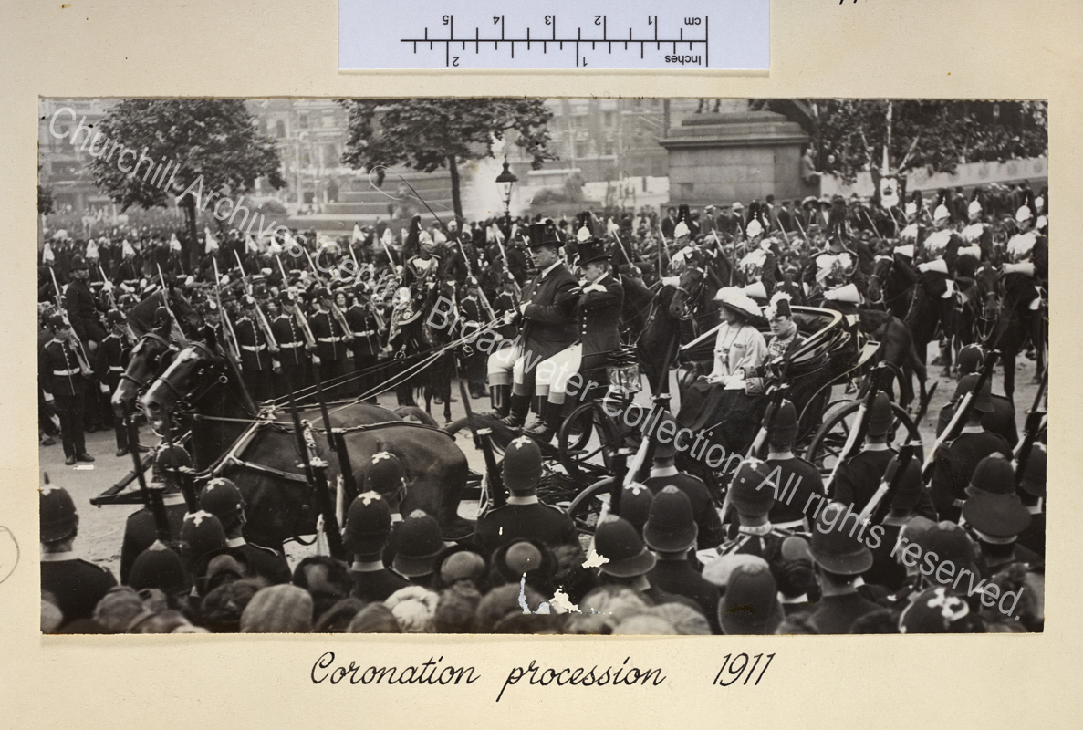 Photo shows WSC and CSC in a horse drawn carriage. WSC is wearing his Privy Councillor's uniform and peaked feathered hat. Police are standing to attention along the route and the carriage is escorted by mounted troops.