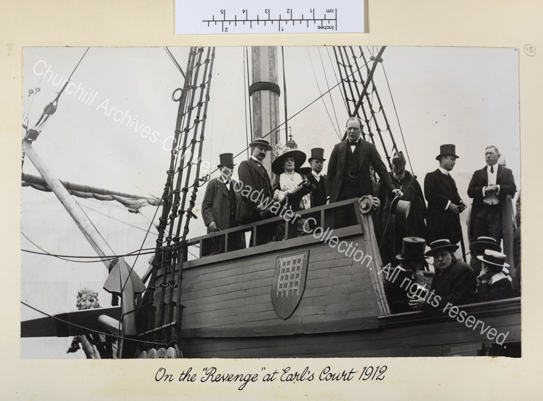 Photo shows WSC holding a top hat and speaking. On the desk with him are several people including Lady Randolph Churchill wearing a wide brimmed hat.