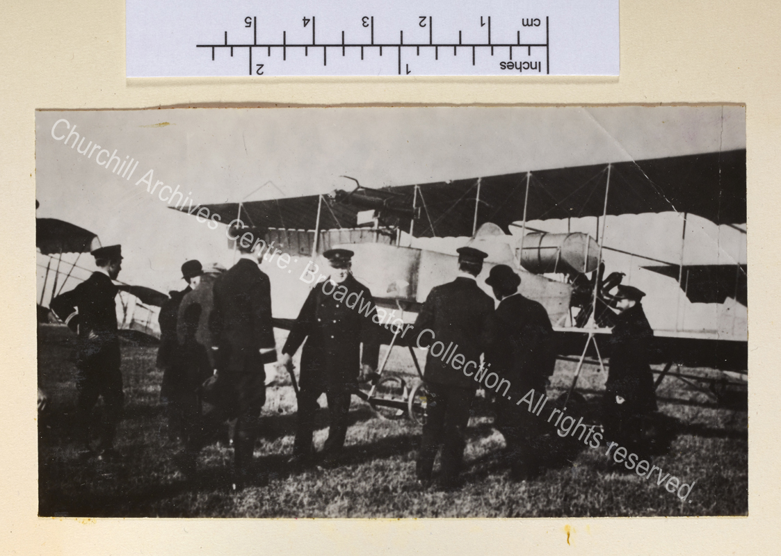 WSC standing in front of a bi-plane wearing an overcoat and peaked cap. WSC is facing towards the camera: others in the group are facing towards the aeroplane.