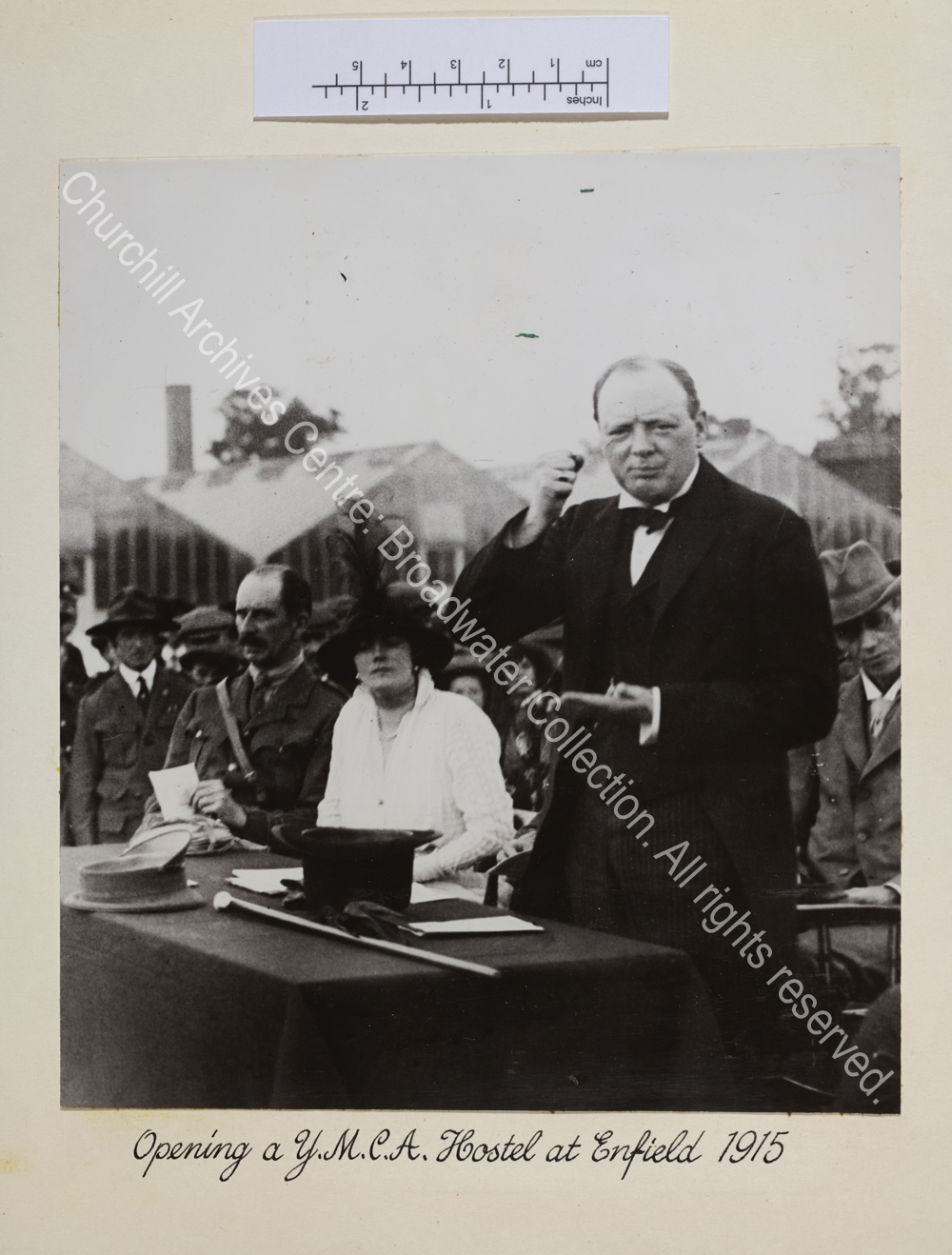 Photo shows WSC standing at a table addressing [munitions workers] using his hands to add emphasis to his words. CSC is seated next to him wearing a hat.