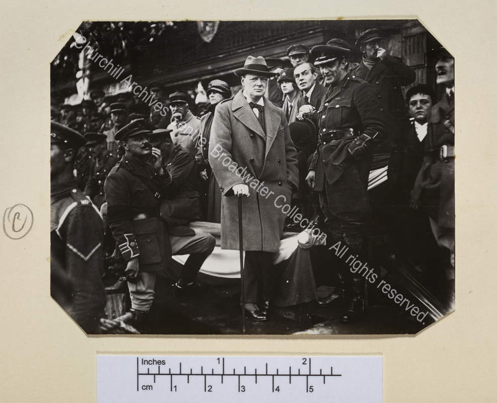 Photo shows WSC standing in a crowd (mainly of men in uniform) leaning on a walking stick. Edward Marsh is to the right of WSC