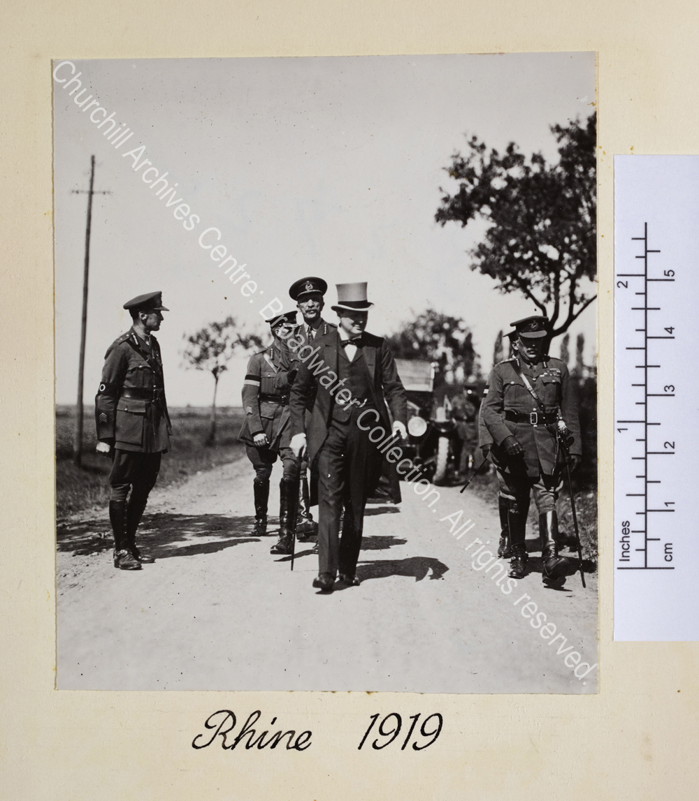 WSC walking along a road wearing top hat and carrying walking stick. To the left