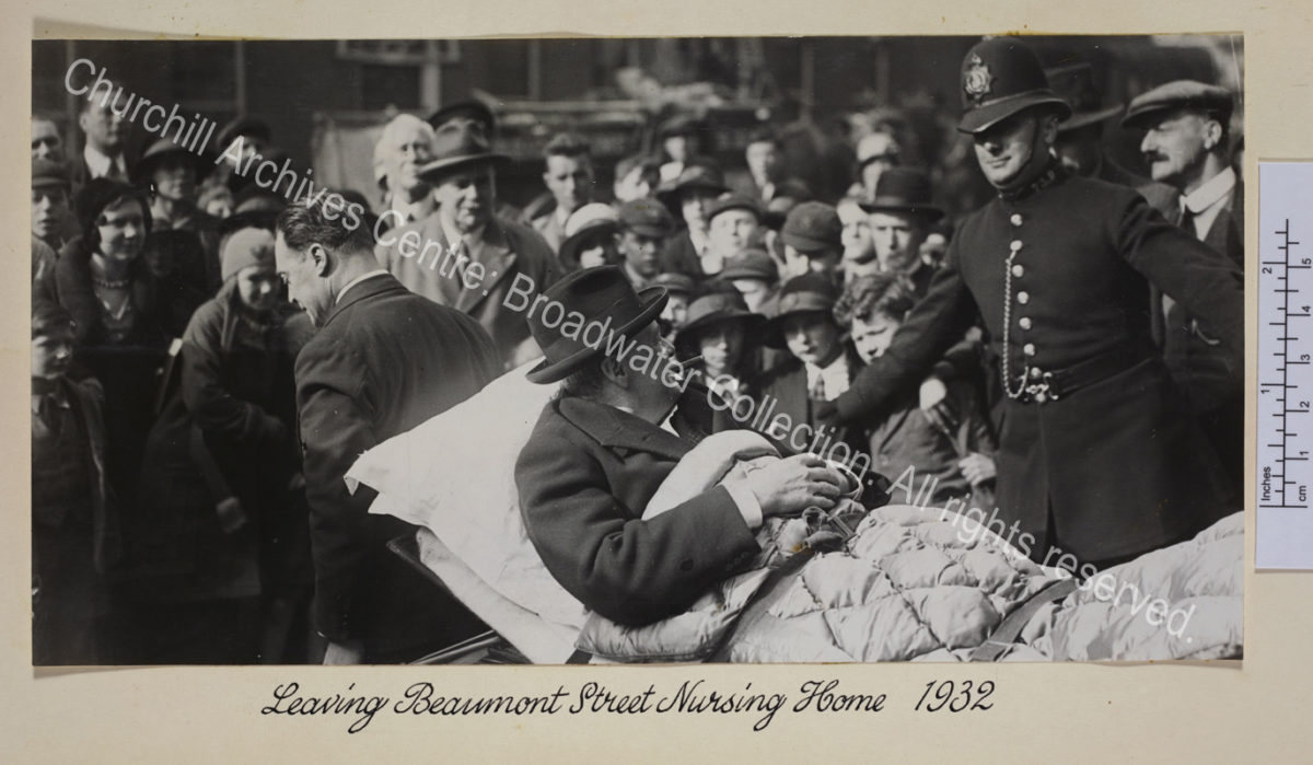 Photo shows WSC on a stretcher being carried through a crowd of people. He is wearing a suit and hat and has a cigar in his mouth.