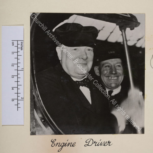 Photo shows WSC smiling and wearing a cap. Just behind him is a man wearing a cap bearing the initials LMS [London