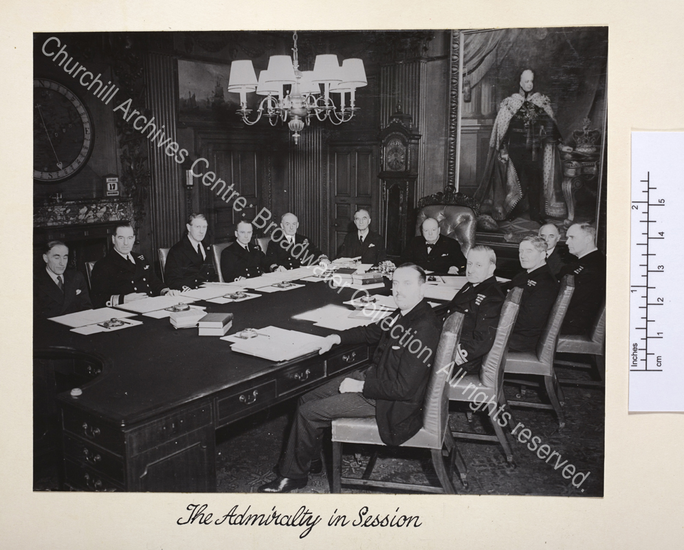Photo showing WSC at the head of a large meeting table. Admiralty staff and officers flank the table with meeting papers and blotters in front of them.