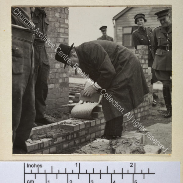 Photo shows WSC bending to lay bricks. He is wearing an overcoat and hat and has a cigar in his mouth. He is observed by a servicemen and a servicewoman in uniform.