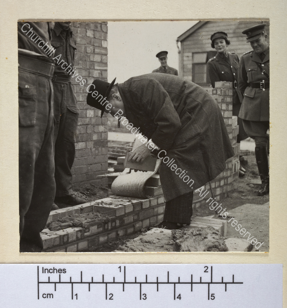 Photo shows WSC bending to lay bricks. He is wearing an overcoat and hat and has a cigar in his mouth. He is observed by a servicemen and a servicewoman in uniform.