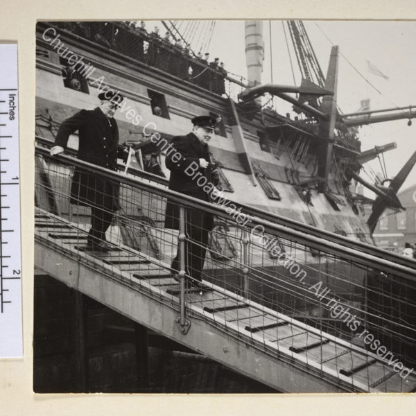 Photo shows WSC wearing an overcoat and peaked cap walking down the gangplank of HMS Victory. Following WSC is Admiral Sir William James