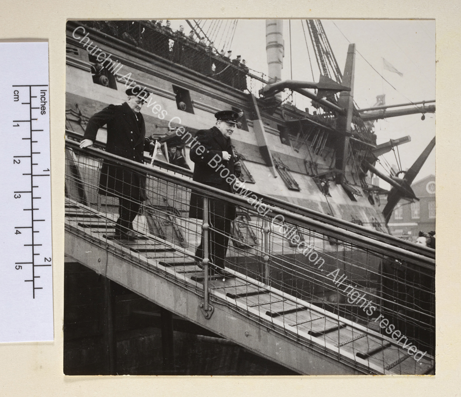 Photo shows WSC wearing an overcoat and peaked cap walking down the gangplank of HMS Victory. Following WSC is Admiral Sir William James