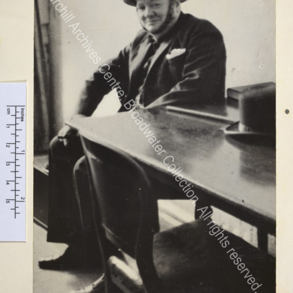 Photo of WSC in Ramsgate [Kent] air raid shelter. Photo shows WSC smiling ruefully wearing a suit and metal helmet. He is sitting next to a table on which rest his Fedora style hat and walking cane.