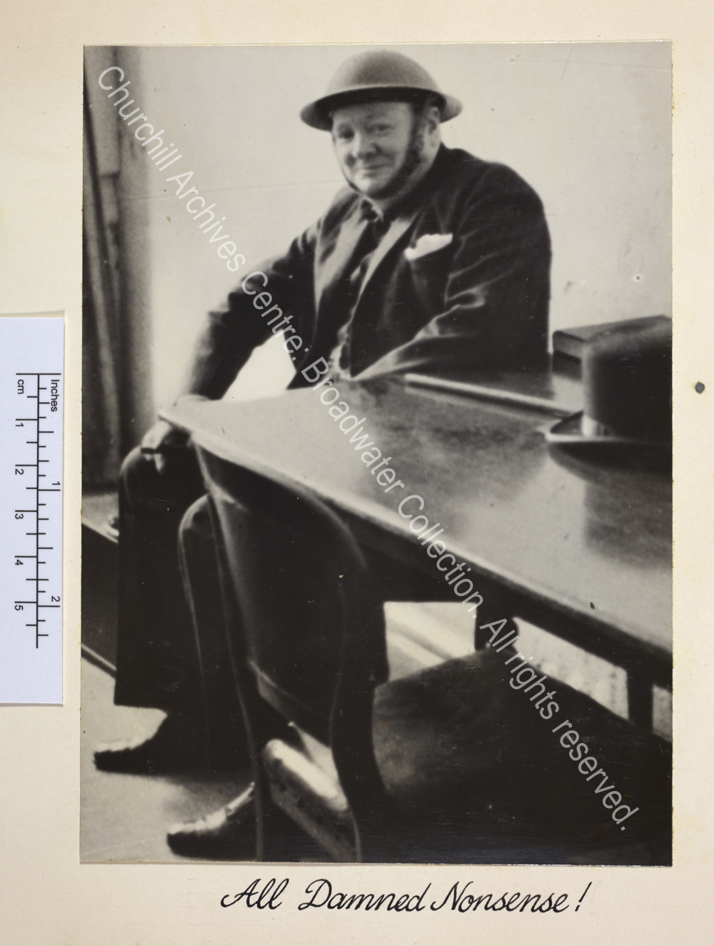 Photo of WSC in Ramsgate [Kent] air raid shelter. Photo shows WSC smiling ruefully wearing a suit and metal helmet. He is sitting next to a table on which rest his Fedora style hat and walking cane.