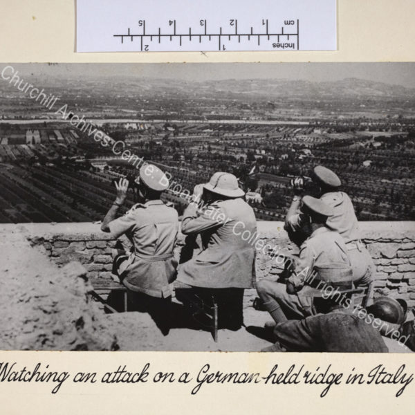 Photo taken during Eighth Army attacks north of Rome. In the foreground is WSC wearing uniform and a pith helmet. He is seated (facing away from the camera) next to the wall of a ruined castle above the front line and looking intently through binoculars. Around him is a group of army officers also watching through binoculars.