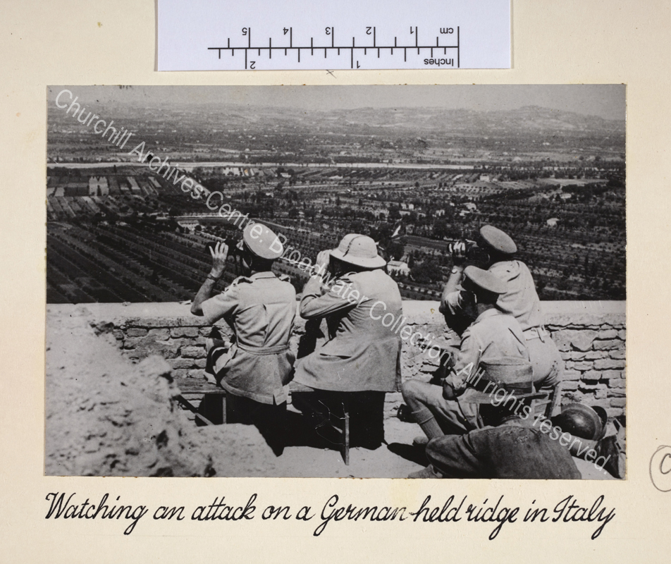Photo taken during Eighth Army attacks north of Rome. In the foreground is WSC wearing uniform and a pith helmet. He is seated (facing away from the camera) next to the wall of a ruined castle above the front line and looking intently through binoculars. Around him is a group of army officers also watching through binoculars.