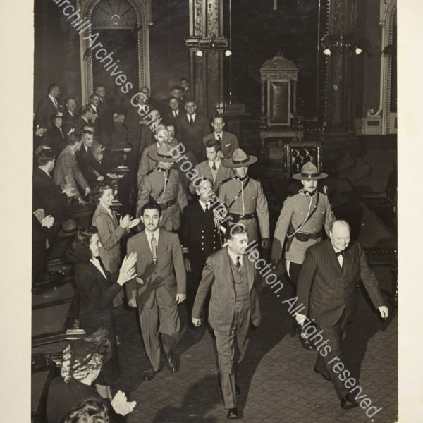 Photo shows WSC's arrival at [?the Canadian Parliament]. He is wearing a pin-striped suit and a bow tie and smiling. He is followed by a group including several Canadian mounted police and is being greeted warmly.