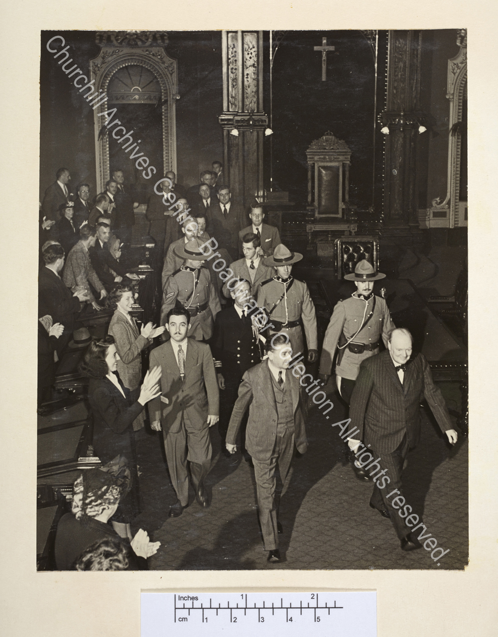 Photo shows WSC's arrival at [?the Canadian Parliament]. He is wearing a pin-striped suit and a bow tie and smiling. He is followed by a group including several Canadian mounted police and is being greeted warmly.