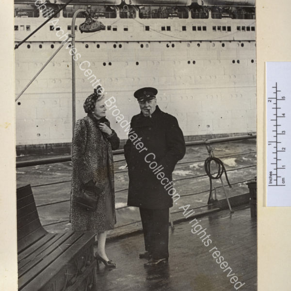 Photo shows WSC and CSC on the deck of the Queen Mary on arrival in Scotland after a visit to Canada. WSC is wearing an overcoat and peaked cap and has a cigar in his mouth and CSC is wearing a fur coat and headscarf.