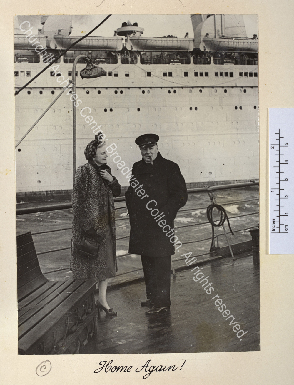 Photo shows WSC and CSC on the deck of the Queen Mary on arrival in Scotland after a visit to Canada. WSC is wearing an overcoat and peaked cap and has a cigar in his mouth and CSC is wearing a fur coat and headscarf.