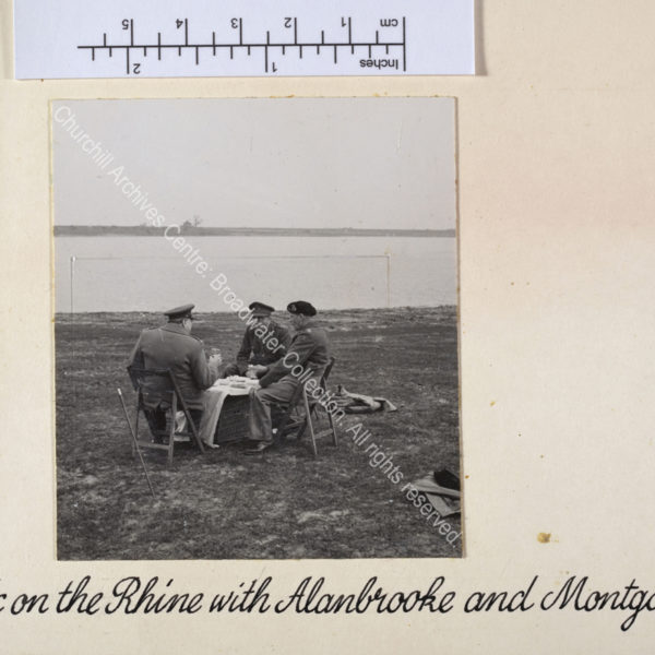 Photo shows WSC (facing away from the camera) seated with Field Marshal Sir Bernard Montgomery [later Lord Montgomery of El Alamein] Commander 21st Army Group and Field Marshal Sir Alan Brooke [later Lord Alanbrooke]. They are all wearing uniform and seated on folding chairs round a wicker hamper covered with a white cloth. Behind them is the river Rhine.