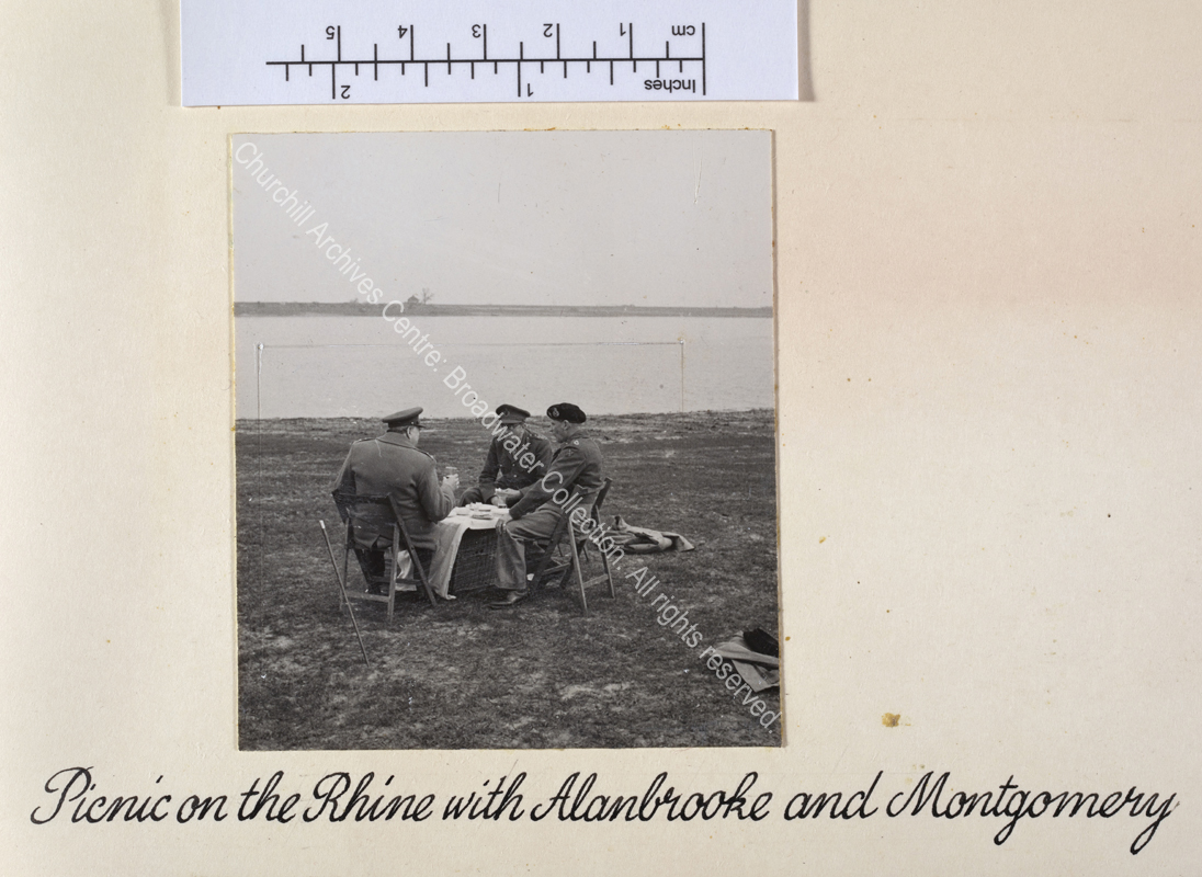Photo shows WSC (facing away from the camera) seated with Field Marshal Sir Bernard Montgomery [later Lord Montgomery of El Alamein] Commander 21st Army Group and Field Marshal Sir Alan Brooke [later Lord Alanbrooke]. They are all wearing uniform and seated on folding chairs round a wicker hamper covered with a white cloth. Behind them is the river Rhine.
