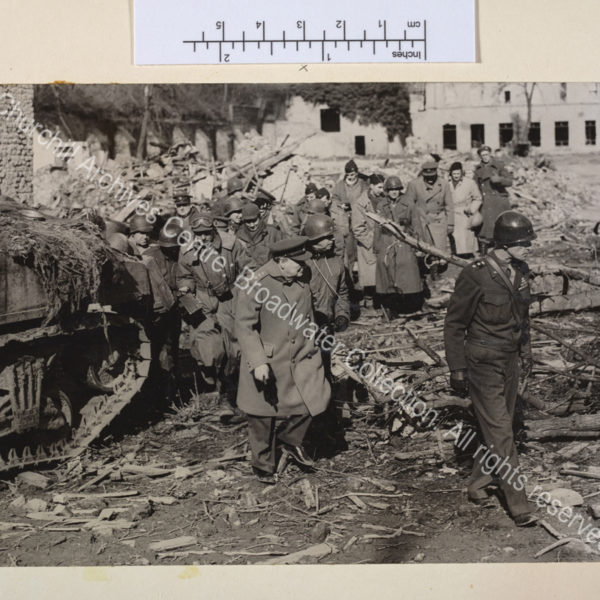 Photo shows WSC in uniform picking his way through rubble and fallen trees followed by a group of people. He is following a US officer in uniform [?General William Simpson of the 9th American Army]. A camouflaged tank is seen at the left of the photo.