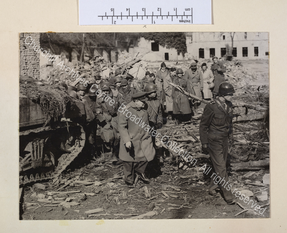Photo shows WSC in uniform picking his way through rubble and fallen trees followed by a group of people. He is following a US officer in uniform [?General William Simpson of the 9th American Army]. A camouflaged tank is seen at the left of the photo.