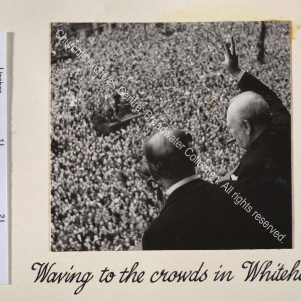 Photo taken from above and behind WSC shows him facing away from the camera and standing at a balcony looking down at a huge crowd of people below. He has a cigar in his mouth and is holding his hand up making a victory sign. A microphone is just seen in front of him.