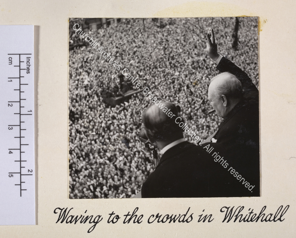 Photo taken from above and behind WSC shows him facing away from the camera and standing at a balcony looking down at a huge crowd of people below. He has a cigar in his mouth and is holding his hand up making a victory sign. A microphone is just seen in front of him.