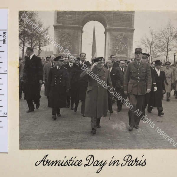 Photo shows WSC and General Charles de Gaulle walking down the Avenue des Champs-Elysee with the Arc de Triomphe behind them during the French Armistice Day parade. Both are in uniform and WSC is raising his hand in salute. Anthony Eden [later 1st Lord Avon] walks behind WSC.