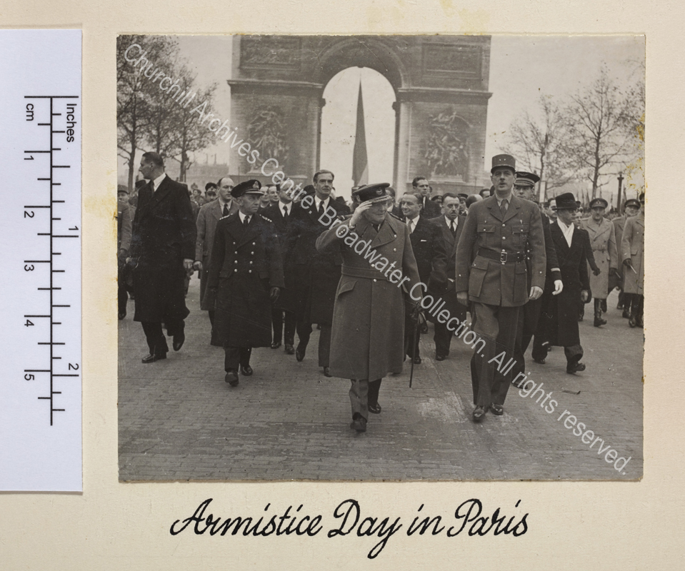 Photo shows WSC and General Charles de Gaulle walking down the Avenue des Champs-Elysee with the Arc de Triomphe behind them during the French Armistice Day parade. Both are in uniform and WSC is raising his hand in salute. Anthony Eden [later 1st Lord Avon] walks behind WSC.