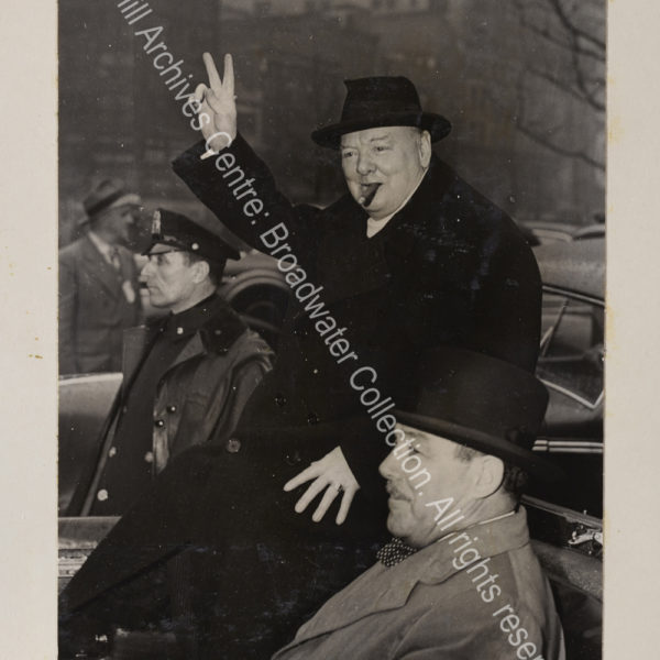 Photo shows WSC in the back of an open topped car on Broadway. He is wearing an overcoat and hat and raising his hand in a victory sign. Grover Whalen sits next to him in the car.