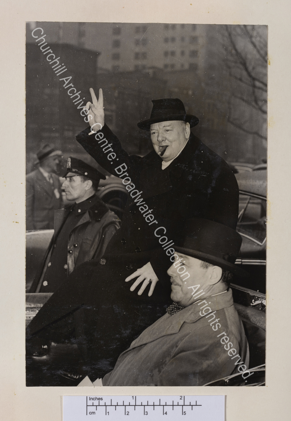 Photo shows WSC in the back of an open topped car on Broadway. He is wearing an overcoat and hat and raising his hand in a victory sign. Grover Whalen sits next to him in the car.